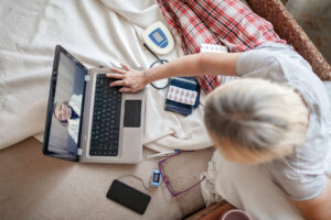 A woman is sitting on her bed, surrounded by a package of pills, a thermometer, and some other medical devices. She is using her laptop to get a virtual second opinion about her rare disease.