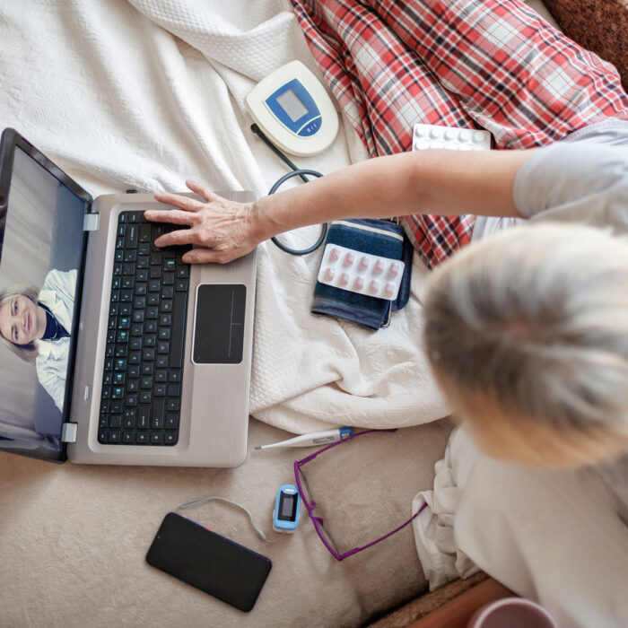 A woman is sitting on her bed, surrounded by a package of pills, a thermometer, and some other medical devices. She is using her laptop to get a virtual second opinion about her rare disease.