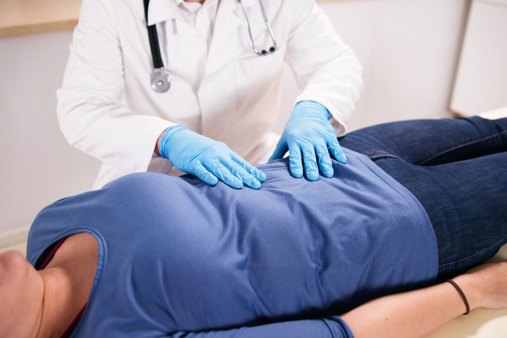 A woman is laying on an exam table, and a physcian is wearing gloves and pressing lightly on her abdomen.